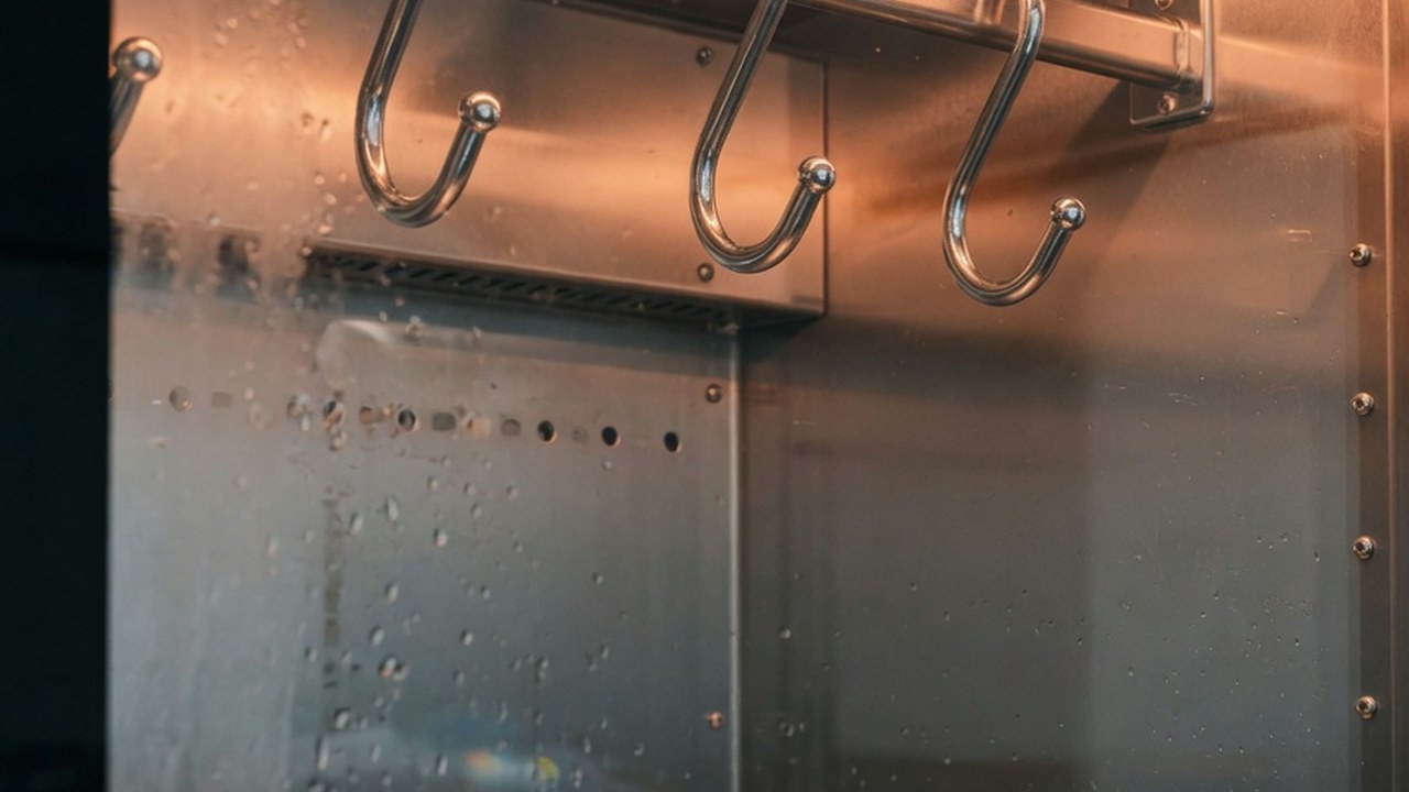 A dry-aging cabinet interior at rest — chrome hooks empty on a stainless rail, warm amber LED strip lighting along the top, a small analog temperature gauge in the corner.